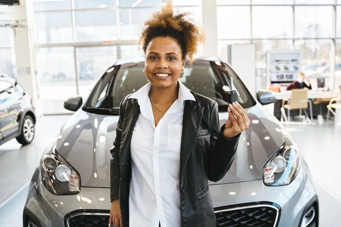 A smiling woman holding car keys beside a car in a showroom.